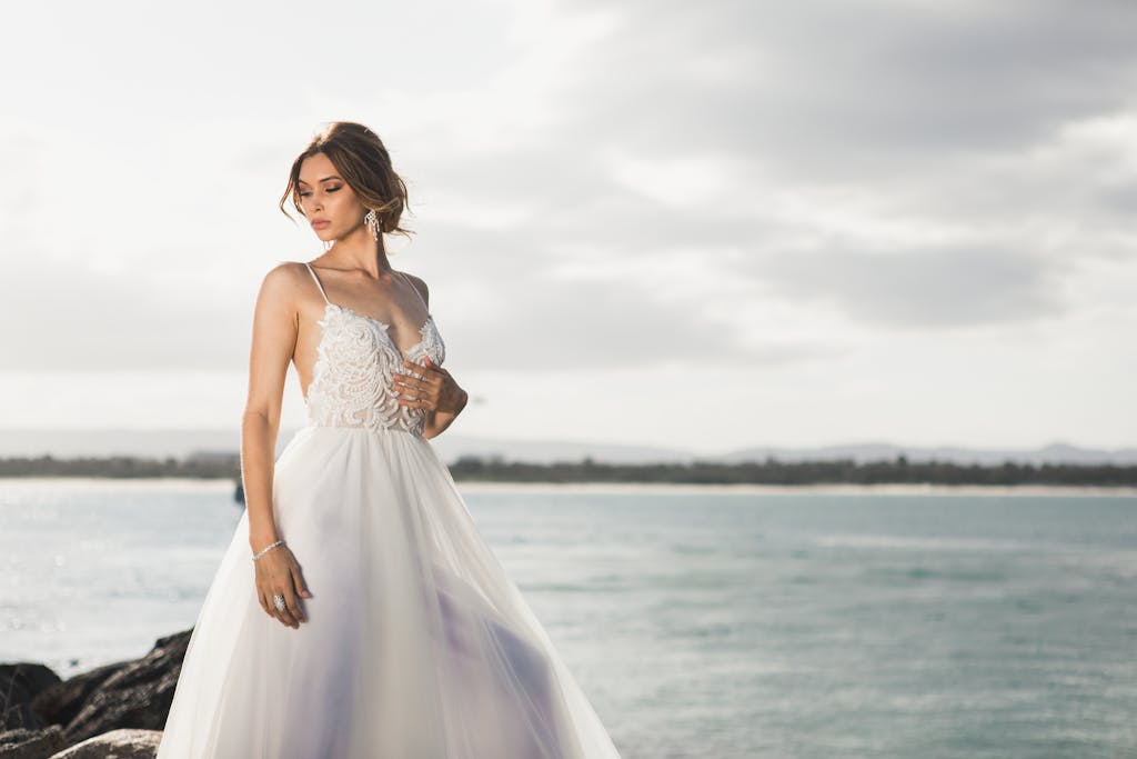 A beautiful bride in a stylish wedding dress posing by the ocean under soft daylight.