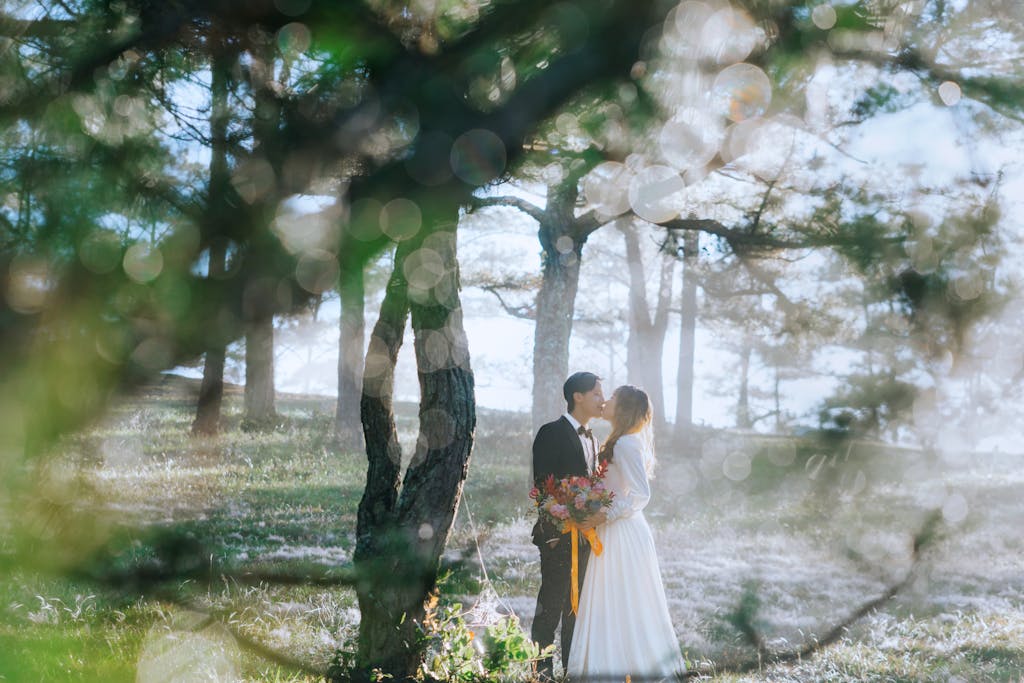 A newlywed couple shares a romantic kiss in a sunlit forest with soft bokeh effects.