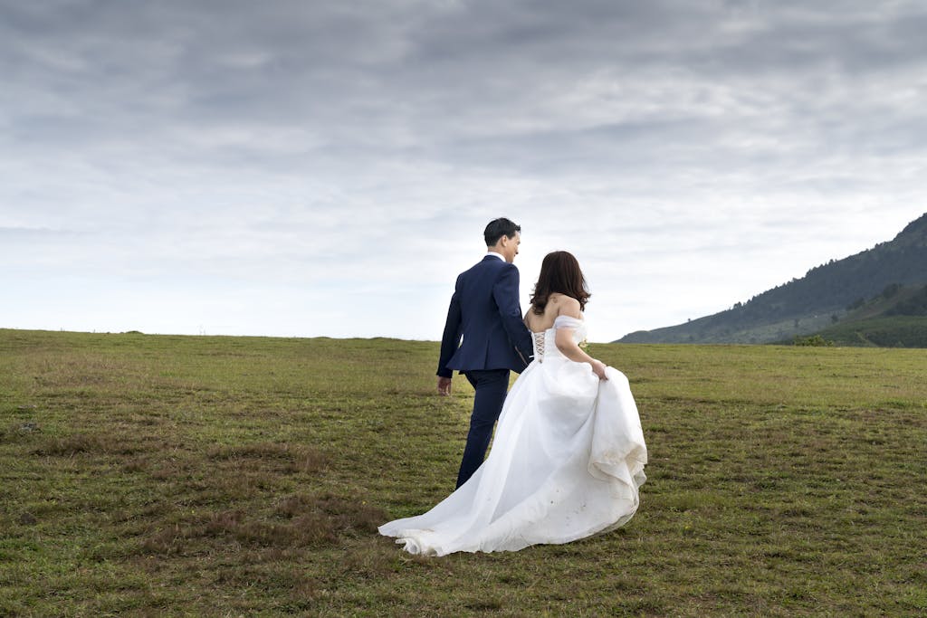 Bride and groom walking in a scenic outdoor field under a cloudy sky, capturing a romantic moment.